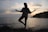 A silhouette of a person in a yoga pose, standing on one leg with arms outstretched, against the backdrop of a serene ocean and a cloudy sky during sunset. The horizon line slightly separates the calm sea from the sky, while darker sections of land line one side of the scene.