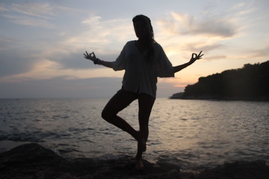 A silhouette of a person in a yoga pose, standing on one leg with arms outstretched, against the backdrop of a serene ocean and a cloudy sky during sunset. The horizon line slightly separates the calm sea from the sky, while darker sections of land line one side of the scene.