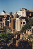 A historical urban scene featuring ancient Roman architecture, including majestic columns, domed roofs, and a backdrop of classical buildings. The structures are composed of stone and brick, set against a clear blue sky. The foreground contains a mix of ruins and lush greenery.