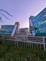 Modern office buildings with glass exteriors tower into the sky, reflecting the evening light. The structures are angled and feature sleek, contemporary designs with visible logos. In the foreground, there is lush greenery and a metal fence.