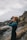 A person wearing a wetsuit and a brown beanie is taking a photo with a camera near a rocky coastline. The sky is overcast, and the ocean is visible, creating a serene and contemplative atmosphere.