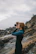 A person wearing a wetsuit and a brown beanie is taking a photo with a camera near a rocky coastline. The sky is overcast, and the ocean is visible, creating a serene and contemplative atmosphere.