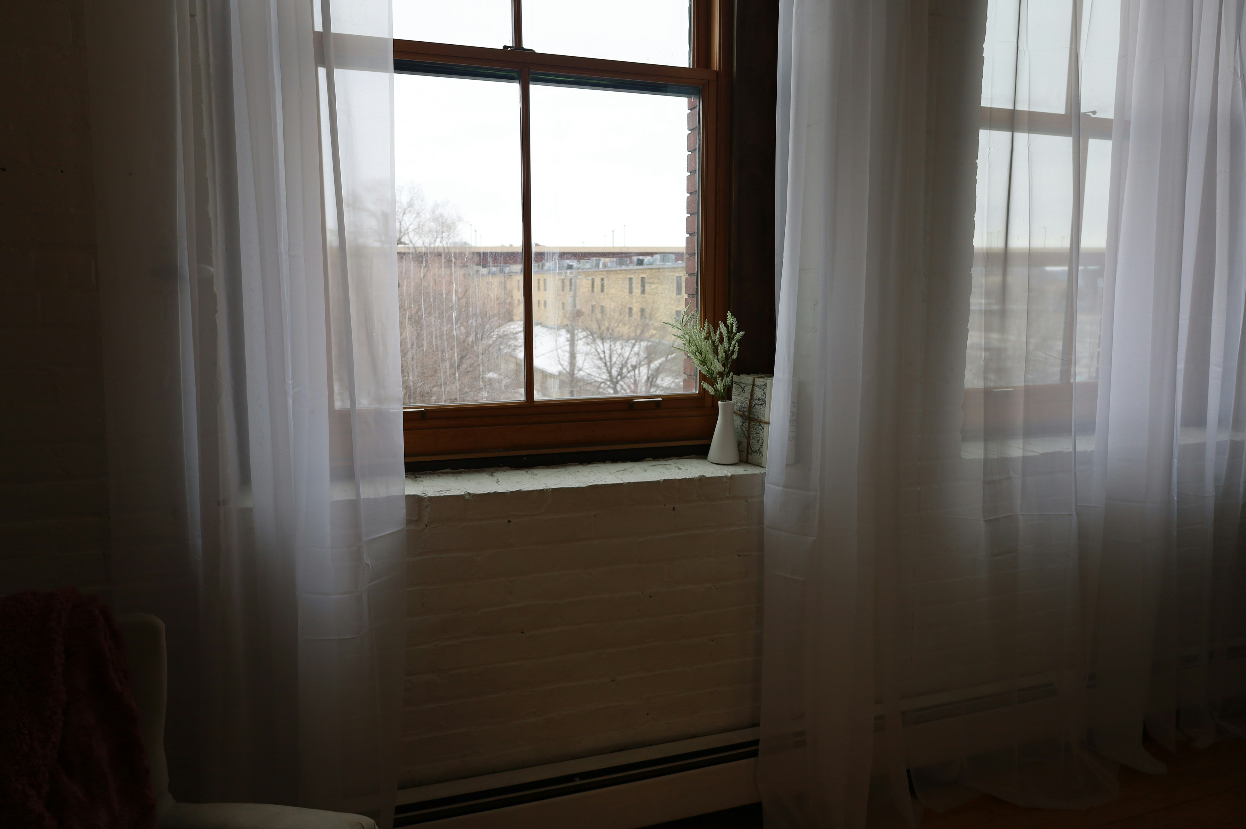 Classic NYC apartment sash window with white sheer curtains, a windowsill vase, and a radiator below