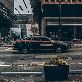 A black police car is parked on a city street, with modern buildings and pedestrians in the background. The street is damp and there are fallen leaves scattered on the ground.