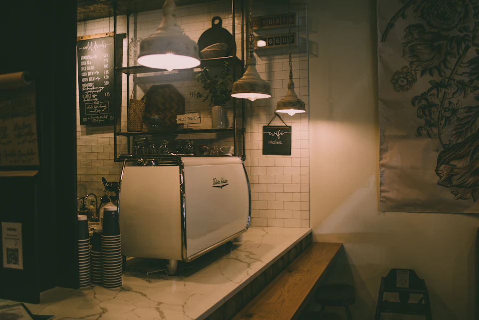 The image depicts a cozy coffee shop interior with white tiled walls and a wooden counter. A white espresso machine is prominently displayed on the counter, surrounded by several cups and saucers arranged neatly around it. Above the counter hangs a blackboard menu, and a sign reading "Coffee" is visible in the background. The scene conveys a sense of warmth and comfort, inviting customers to enjoy their coffee experience.
