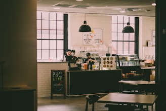 A cozy café setting featuring large windows and industrial-style pendant lights. The counter is attended by staff members, and the menu is displayed above. A board near the counter reads 'PHO LIFE', with a small illustration of a bowl of pho. The interior has a warm ambiance with wooden furniture and white-tiled walls.