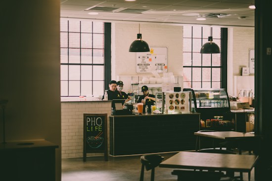 A cozy caf&eacute; setting featuring large windows and industrial-style pendant lights. The counter is attended by staff members, and the menu is displayed above. A board near the counter reads 'PHO LIFE', with a small illustration of a bowl of pho. The interior has a warm ambiance with wooden furniture and white-tiled walls.