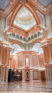 An intricately designed interior features geometric patterns, archways, and a domed ceiling adorned with detailed mosaics and carvings. The space exhibits rich architectural elements including stained glass windows and polished marble surfaces.