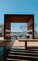 A wooden pergola frames a view of a distant shoreline across a body of water. Shadows from the structure create patterns on the ground. Several trees can be seen near the pergola and in the distance. The sky is clear and blue, enhancing the serene atmosphere.
