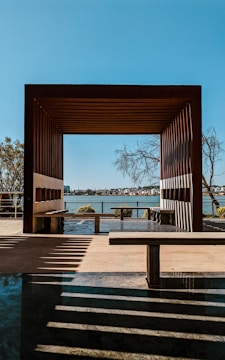 A wooden pergola frames a view of a distant shoreline across a body of water. Shadows from the structure create patterns on the ground. Several trees can be seen near the pergola and in the distance. The sky is clear and blue, enhancing the serene atmosphere.