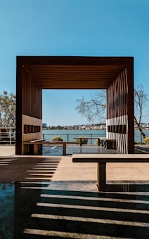 A wooden pergola frames a view of a distant shoreline across a body of water. Shadows from the structure create patterns on the ground. Several trees can be seen near the pergola and in the distance. The sky is clear and blue, enhancing the serene atmosphere.