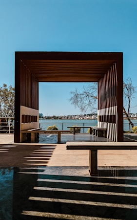 A wooden pergola frames a view of a distant shoreline across a body of water. Shadows from the structure create patterns on the ground. Several trees can be seen near the pergola and in the distance. The sky is clear and blue, enhancing the serene atmosphere.