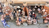 A street vendor is selling a variety of copper goods, including pots, pans, and decorative items. The shop is outdoors with an open entrance, and various copper items are hanging and displayed on tables. The vendor, dressed in casual clothing, is working amidst the merchandise. Dry leaves and branches are visible in the foreground, indicating an autumn setting.