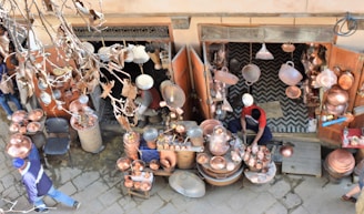 A street vendor is selling a variety of copper goods, including pots, pans, and decorative items. The shop is outdoors with an open entrance, and various copper items are hanging and displayed on tables. The vendor, dressed in casual clothing, is working amidst the merchandise. Dry leaves and branches are visible in the foreground, indicating an autumn setting.