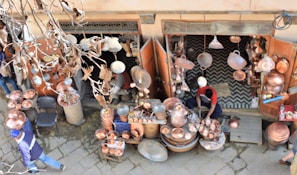 A street vendor is selling a variety of copper goods, including pots, pans, and decorative items. The shop is outdoors with an open entrance, and various copper items are hanging and displayed on tables. The vendor, dressed in casual clothing, is working amidst the merchandise. Dry leaves and branches are visible in the foreground, indicating an autumn setting.