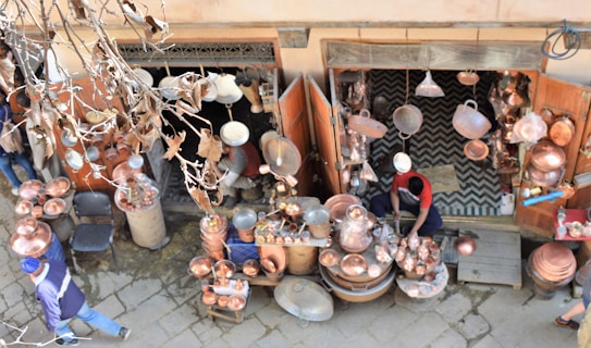 A street vendor is selling a variety of copper goods, including pots, pans, and decorative items. The shop is outdoors with an open entrance, and various copper items are hanging and displayed on tables. The vendor, dressed in casual clothing, is working amidst the merchandise. Dry leaves and branches are visible in the foreground, indicating an autumn setting.