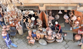 A street vendor is selling a variety of copper goods, including pots, pans, and decorative items. The shop is outdoors with an open entrance, and various copper items are hanging and displayed on tables. The vendor, dressed in casual clothing, is working amidst the merchandise. Dry leaves and branches are visible in the foreground, indicating an autumn setting.