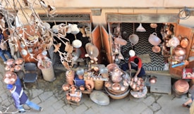 A street vendor is selling a variety of copper goods, including pots, pans, and decorative items. The shop is outdoors with an open entrance, and various copper items are hanging and displayed on tables. The vendor, dressed in casual clothing, is working amidst the merchandise. Dry leaves and branches are visible in the foreground, indicating an autumn setting.
