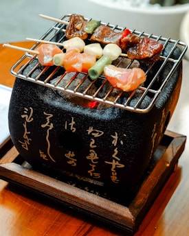 Skewers of meat and vegetables are placed on a small charcoal grill with a black ceramic body featuring Japanese script. The wooden base adds a traditional touch to the setup.