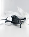 A black quadcopter drone with four propellers is placed on white crumpled paper. The drone features a camera at its front and has red and silver accents on its propellers. The background is a soft white, creating a clean and minimalistic setting.