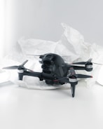 A black quadcopter drone with four propellers is placed on white crumpled paper. The drone features a camera at its front and has red and silver accents on its propellers. The background is a soft white, creating a clean and minimalistic setting.