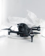 A black quadcopter drone with four propellers is placed on white crumpled paper. The drone features a camera at its front and has red and silver accents on its propellers. The background is a soft white, creating a clean and minimalistic setting.