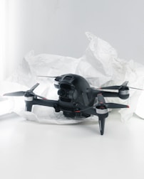 A black quadcopter drone with four propellers is placed on white crumpled paper. The drone features a camera at its front and has red and silver accents on its propellers. The background is a soft white, creating a clean and minimalistic setting.