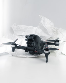 A black quadcopter drone with four propellers is placed on white crumpled paper. The drone features a camera at its front and has red and silver accents on its propellers. The background is a soft white, creating a clean and minimalistic setting.