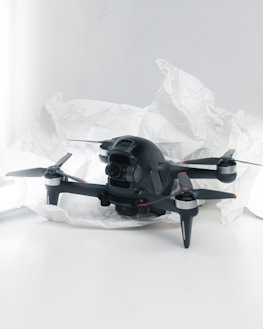 A black quadcopter drone with four propellers is placed on white crumpled paper. The drone features a camera at its front and has red and silver accents on its propellers. The background is a soft white, creating a clean and minimalistic setting.