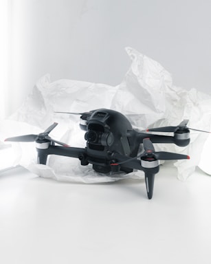 A black quadcopter drone with four propellers is placed on white crumpled paper. The drone features a camera at its front and has red and silver accents on its propellers. The background is a soft white, creating a clean and minimalistic setting.