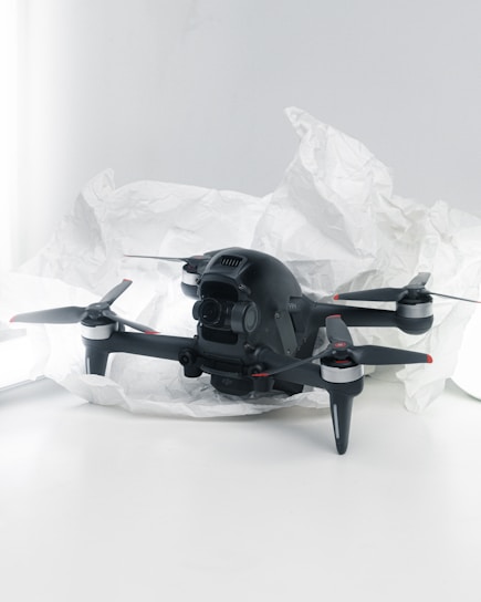 A black quadcopter drone with four propellers is placed on white crumpled paper. The drone features a camera at its front and has red and silver accents on its propellers. The background is a soft white, creating a clean and minimalistic setting.