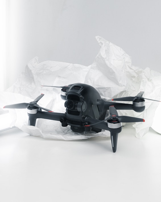 A black quadcopter drone with four propellers is placed on white crumpled paper. The drone features a camera at its front and has red and silver accents on its propellers. The background is a soft white, creating a clean and minimalistic setting.