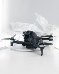 A black quadcopter drone with four propellers is placed on white crumpled paper. The drone features a camera at its front and has red and silver accents on its propellers. The background is a soft white, creating a clean and minimalistic setting.