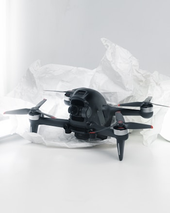 A black quadcopter drone with four propellers is placed on white crumpled paper. The drone features a camera at its front and has red and silver accents on its propellers. The background is a soft white, creating a clean and minimalistic setting.