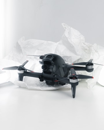 A black quadcopter drone with four propellers is placed on white crumpled paper. The drone features a camera at its front and has red and silver accents on its propellers. The background is a soft white, creating a clean and minimalistic setting.