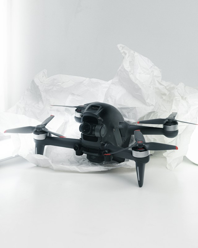 A black quadcopter drone with four propellers is placed on white crumpled paper. The drone features a camera at its front and has red and silver accents on its propellers. The background is a soft white, creating a clean and minimalistic setting.