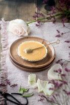 A rustic tabletop setting featuring a small round dish filled with honey on a wooden plate, accompanied by a honey dipper. Surrounding the dish are delicate pink rose petals and a full rose. Lush sprigs of purple flowers add a touch of color, contrasting against the light fabric covering the wooden surface.