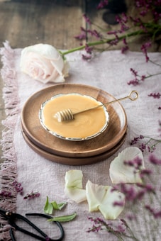 A rustic tabletop setting featuring a small round dish filled with honey on a wooden plate, accompanied by a honey dipper. Surrounding the dish are delicate pink rose petals and a full rose. Lush sprigs of purple flowers add a touch of color, contrasting against the light fabric covering the wooden surface.