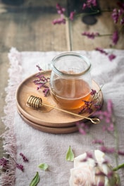A jar filled with honey sits on a wooden plate accompanied by a honey dipper. The scene is adorned with sprigs of purple flowers and a delicate rose, all set atop a textured fabric tablecloth.