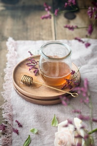 A jar filled with honey sits on a wooden plate accompanied by a honey dipper. The scene is adorned with sprigs of purple flowers and a delicate rose, all set atop a textured fabric tablecloth.
