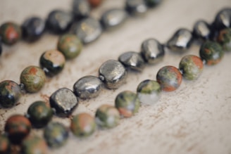 A close-up view of a string of beads with a variety of colors and textures. The beads have a natural, earthy appearance with hues of green, brown, and metallic tones. The background is a light, neutral color that highlights the beads.