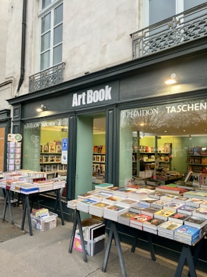 A bookshop named 'Art Book' has a display window showing various art books and magazines. Outside, tables are filled with stacks of books, arranged neatly. The shop facade is adorned with ornate ironwork railing above the windows.