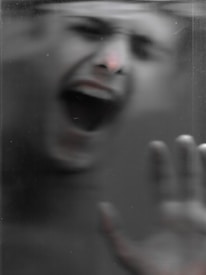 A blurred, grayscale image of a person with their mouth wide open and one hand pressed against a surface, possibly evoking a sense of distress or urgency.
