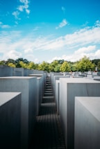 Tall concrete slabs arranged in a grid pattern form a large outdoor installation. The slabs are placed on an uneven ground surface, creating various heights and walkways between them. In the background, green trees and a bright blue sky with scattered clouds add contrast to the otherwise monochromatic structure.