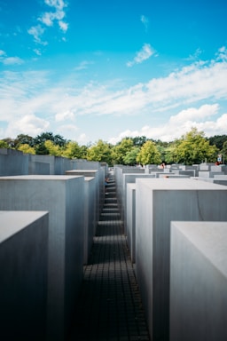 Tall concrete slabs arranged in a grid pattern form a large outdoor installation. The slabs are placed on an uneven ground surface, creating various heights and walkways between them. In the background, green trees and a bright blue sky with scattered clouds add contrast to the otherwise monochromatic structure.