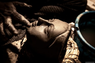 A hand is carefully sculpting a clay face, capturing fine details and expressions. The setting appears dimly lit, with shadows enhancing the intricate textures of the clay.