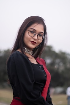 A young woman with long dark hair wearing glasses is dressed in a black blouse and red sari. She accessorizes with a choker necklace. The background is an outdoor setting with blurred greenery.