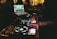 A DJ booth with a laptop displaying music tracks, and DJ equipment with various buttons and turntables. The scene is dimly lit, suggesting a nighttime or club environment with blurred figures in the background and bottles lined up on shelves.