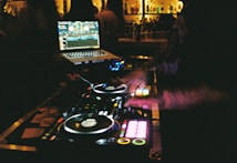 A DJ booth with a laptop displaying music tracks, and DJ equipment with various buttons and turntables. The scene is dimly lit, suggesting a nighttime or club environment with blurred figures in the background and bottles lined up on shelves.
