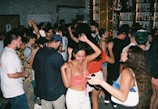 A lively scene at a crowded party with people dancing and socializing. The dimly lit room is adorned with colorful lights, and the background features shelves filled with various bottles. The attendees are dressed casually, enjoying the energetic atmosphere.
